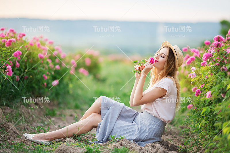 美丽的年轻女孩戴着草帽坐在花园里，周围有粉红色的玫瑰