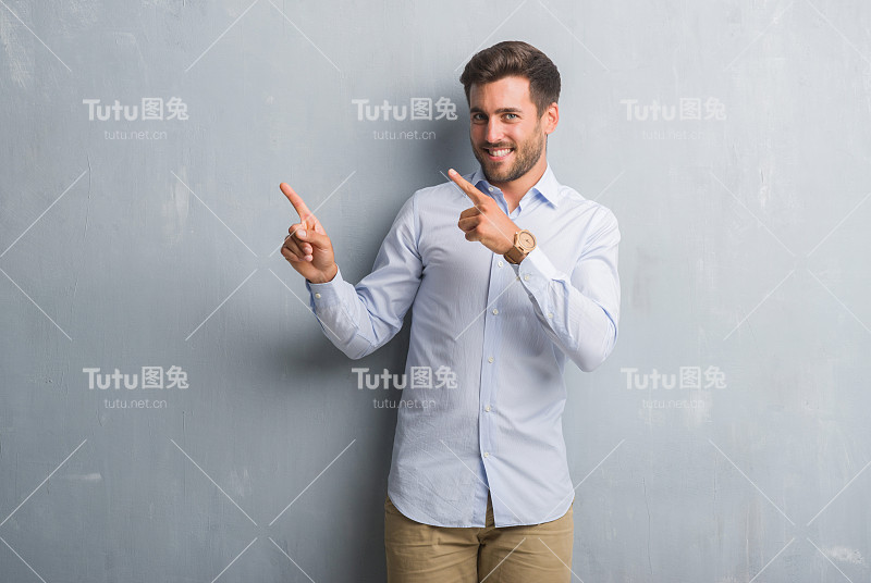 英俊的年轻商人越过灰色垃圾墙，穿着优雅的衬衫，微笑着看着镜头，双手和手指指向一边。