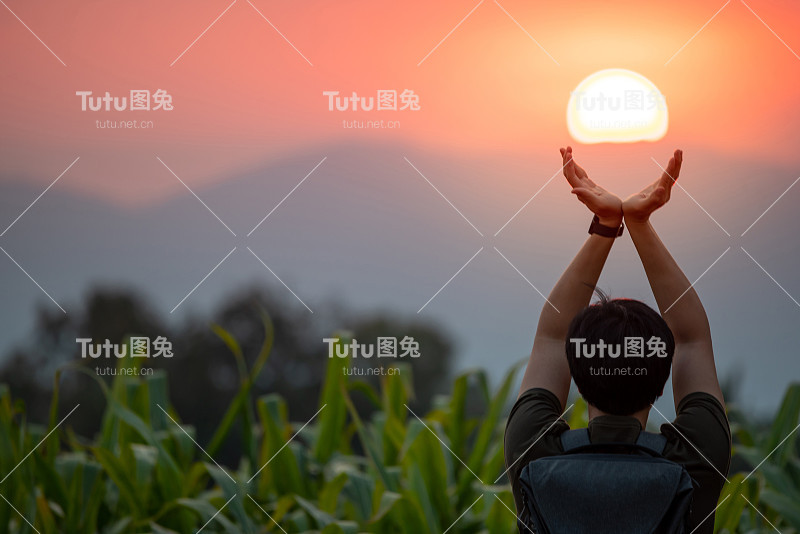 一名亚洲男子在玉米地里举起双臂，在日落时捧着太阳。希望和祈祷。成就的概念