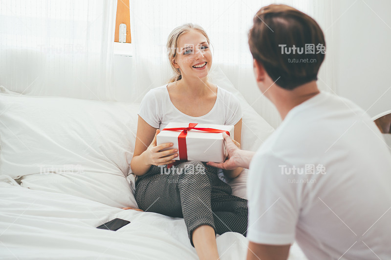 幸福的白人夫妇的爱是在家里的卧室一起庆祝，年轻英俊的男子给他的女朋友礼物盒在情人节，周年纪念，生日或圣诞节惊喜