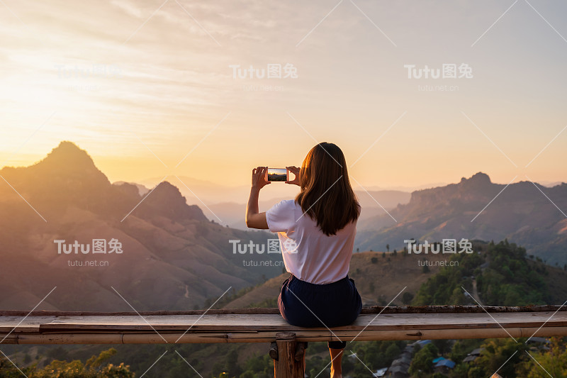 年轻的女人旅行者在日落时用智能手机拍照的山