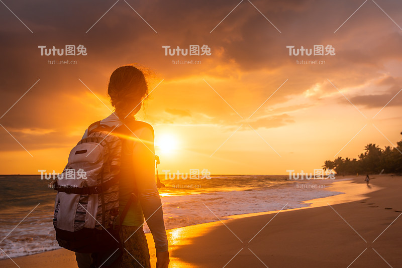 暑假里背着背包的女孩在美丽的日落背景下沿着海岸。