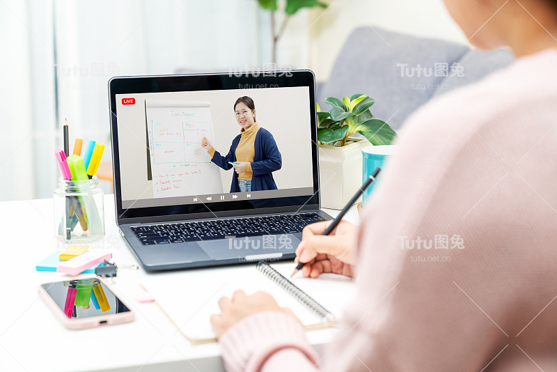 后视图的年轻亚洲女孩坐在桌子上学习在家里的客厅远程学习的概念，虚拟在线，自我隔离隔离的生活方式在亚洲。在电脑上进行视频教学。