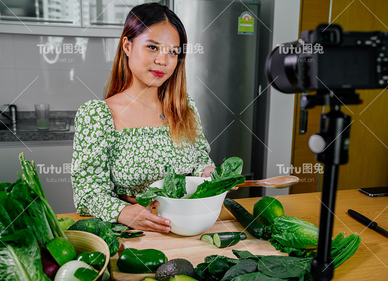 亚洲女性博主或内容创作者烹饪和录制摄像机。在厨房用摄像机拍摄的同时展示健康食品