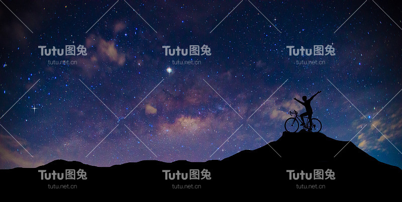 山顶上骑自行车的人的轮廓。全景，蓝色的夜空，银河和星星在黑暗的背景。宇宙充满了恒星，星云和星系的噪声和颗粒。