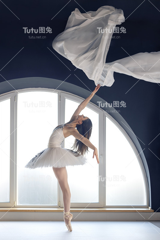 年轻上进的芭蕾舞女演员身穿白色芭蕾舞裙，在自然的日光环境下，在巨大的圆形窗背景前练习芭蕾舞动作