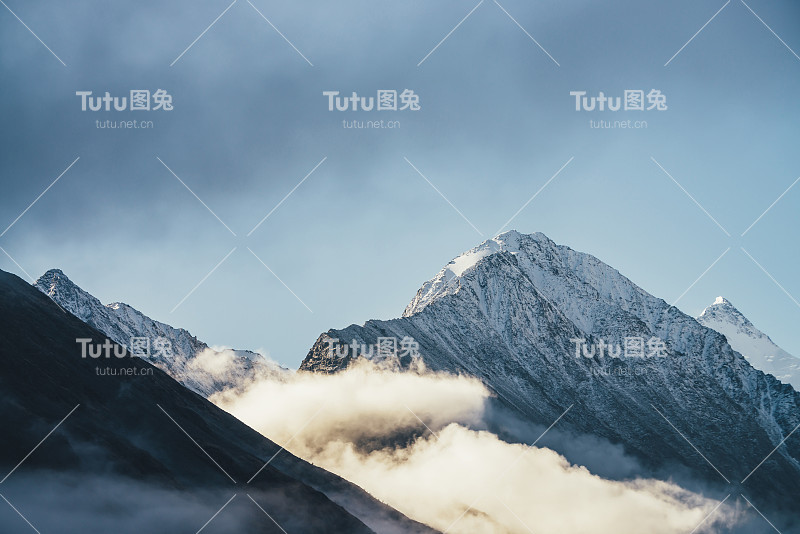 美丽的雪山在阳光下厚厚的云层之上。风景明亮的高山景观，白雪皑皑的山峰在浓密的低云蓝天中。美丽的风景与雪峰。