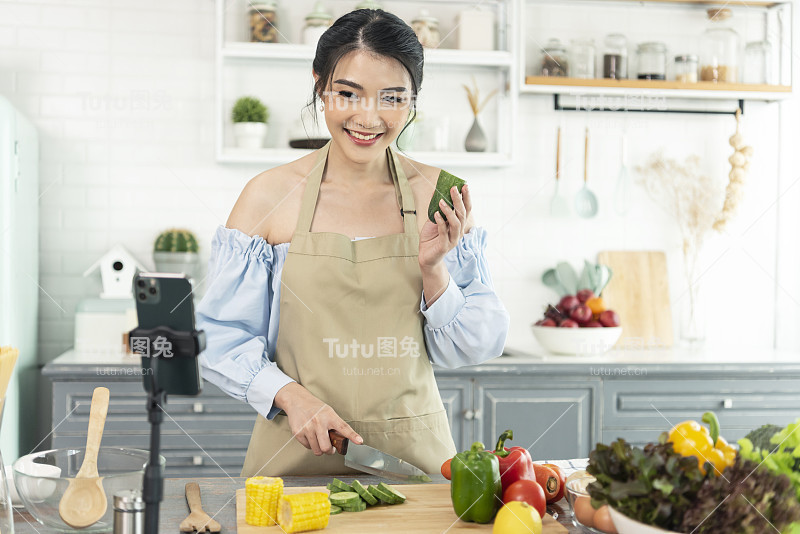 亚洲女性美食博主在智能手机摄像头前烹饪沙拉，同时在家里的厨房录制vlog视频和直播。