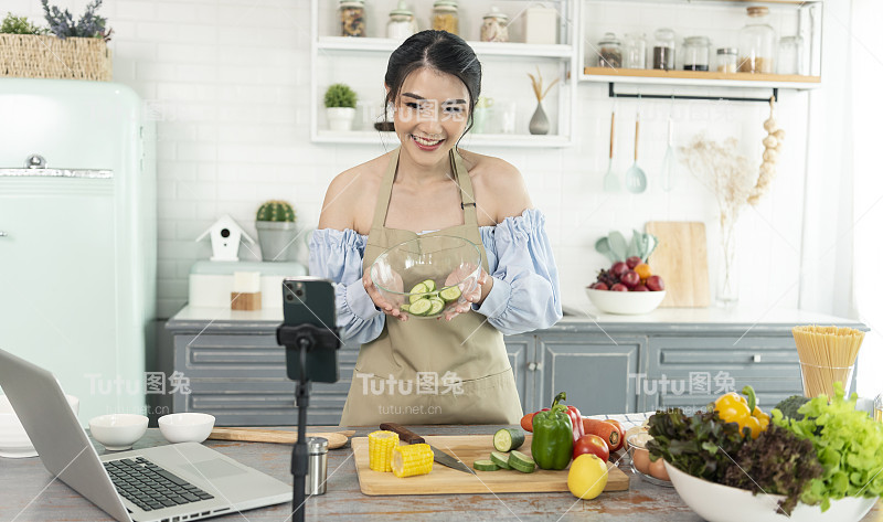 亚洲女性美食博主在智能手机摄像头前烹饪沙拉，同时在家里的厨房录制vlog视频和直播。