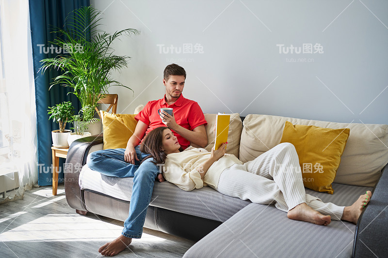 一对美丽的年轻夫妇坐在沙发上，一个女人把头靠在一个男人的膝盖上看书。关系，温柔，爱，和谐的夫妻