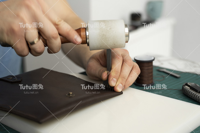 一名男子的手拿着皮革雕刻锤和钢凿孔打孔，在他的车间里制作一个皮革钱包。工作过程中使用棕色天然皮革。手拿工具的工匠。