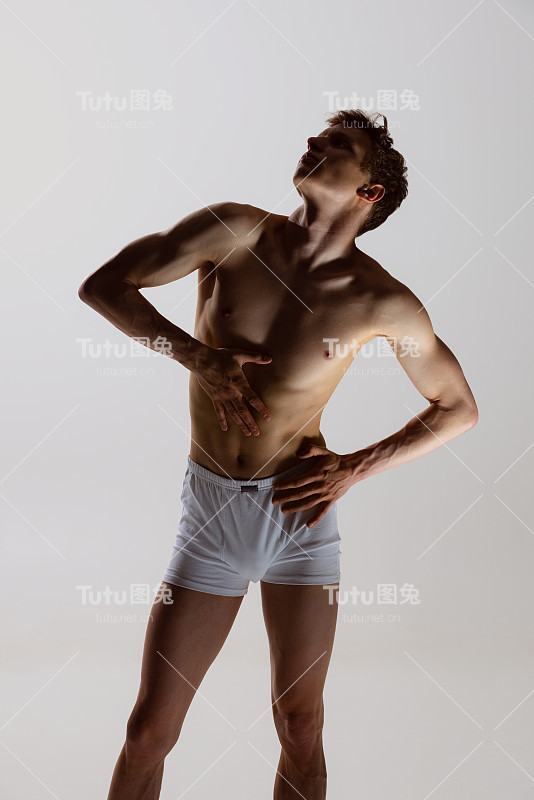 一个赤裸上身、肌肉发达的年轻人的剪影，穿着白色的四角裤，孤立地站在灰色的背景上。男性身体的自然美。情感,爱,关心